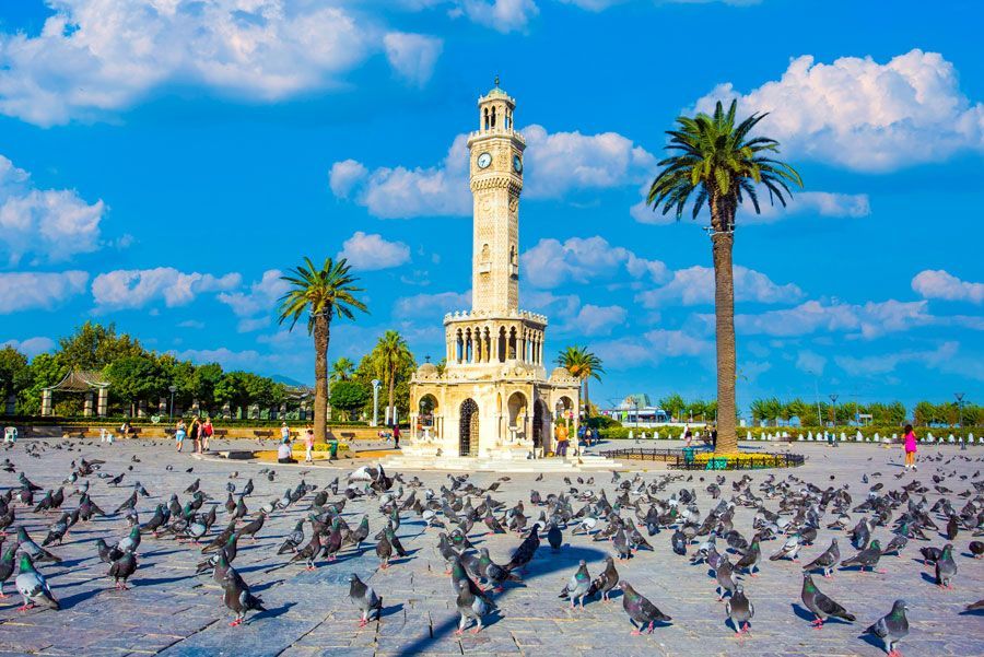 Torre del Reloj de Esmirna, Turquía, con palomas en la plaza bajo un cielo azul. Palmeras flanquean la torre.