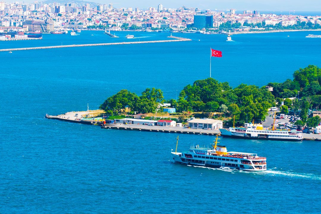 Un ferry navega por aguas azules hacia una pequeña isla con bandera turca ondeando. El horizonte de la ciudad al fondo.