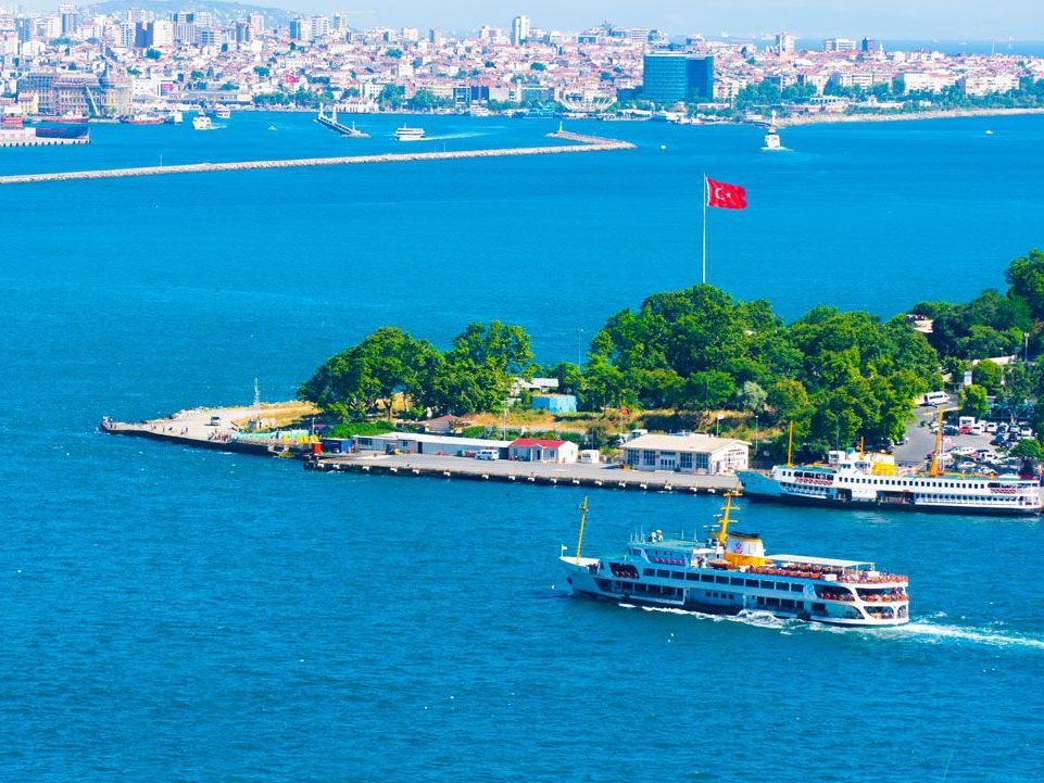 Un ferry navega por aguas azules hacia una isla con una gran bandera turca ondeando. El horizonte de la ciudad al fondo.