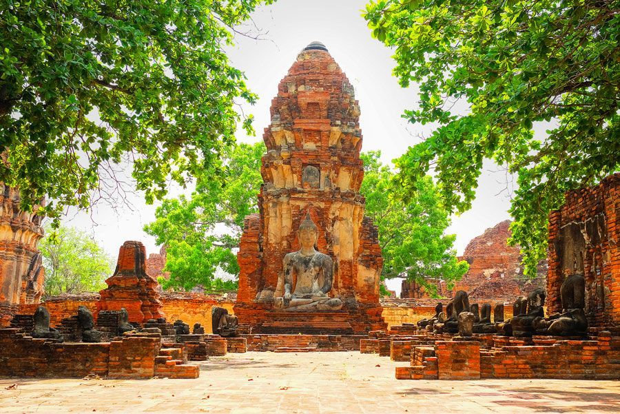 Ruinas de un templo de ladrillo en Tailandia, enmarcadas por frondosos árboles. Una pagoda central alberga una estatua de Buda sedente.