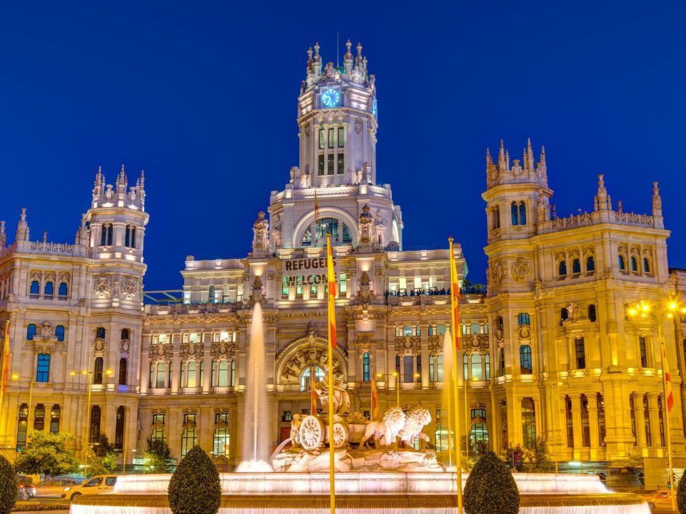 Vista nocturna del Palacio de Cibeles en Madrid, España, iluminado con luces blancas contra un cielo azul oscuro, con una fuente al frente.