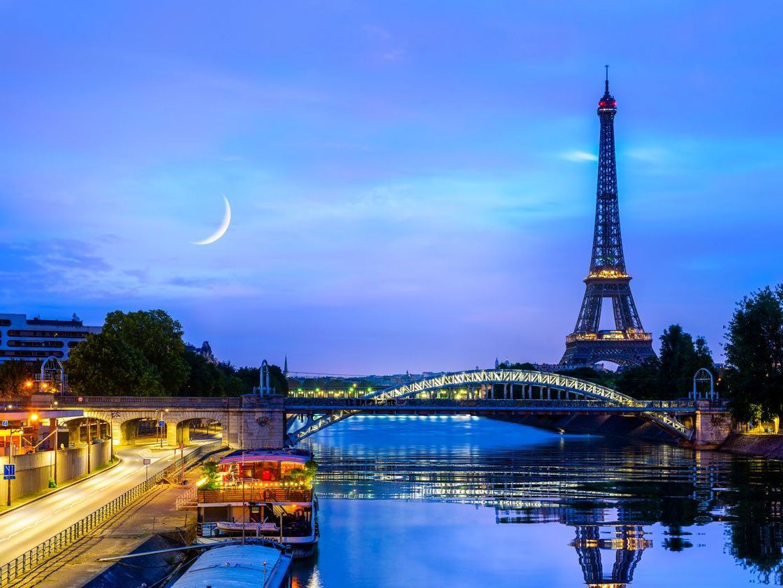 Torre Eiffel y puente sobre un río reflejándose en el agua al anochecer, con una luna creciente en el cielo.