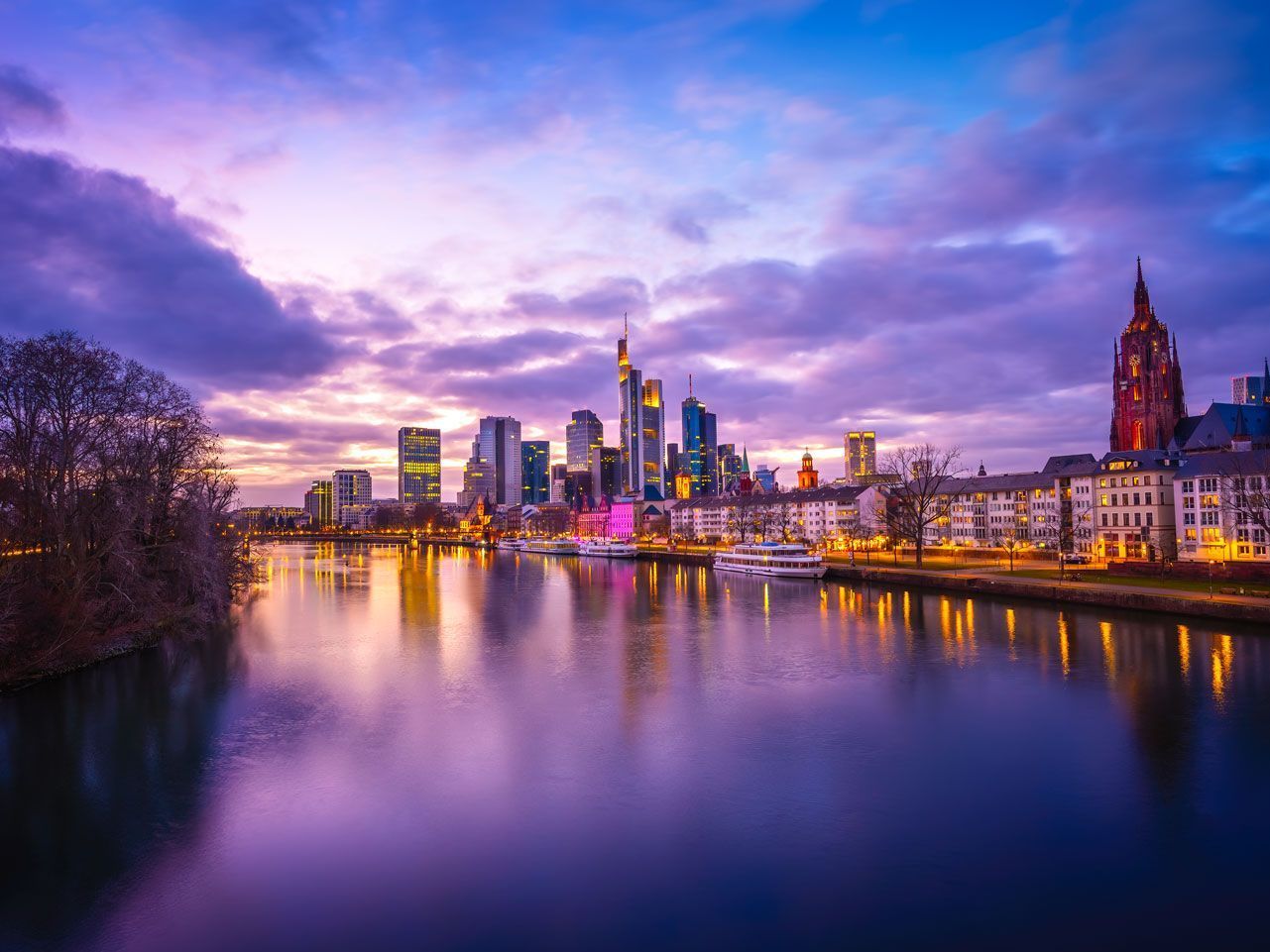 El horizonte de Fráncfort, Alemania, al anochecer, se refleja en el río Meno. Los edificios se recortan contra un cielo morado y naranja.