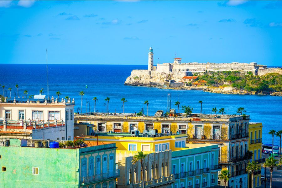 Edificios coloridos con vistas al océano en La Habana, Cuba, con un faro y una fortaleza en la distancia bajo un cielo azul claro.