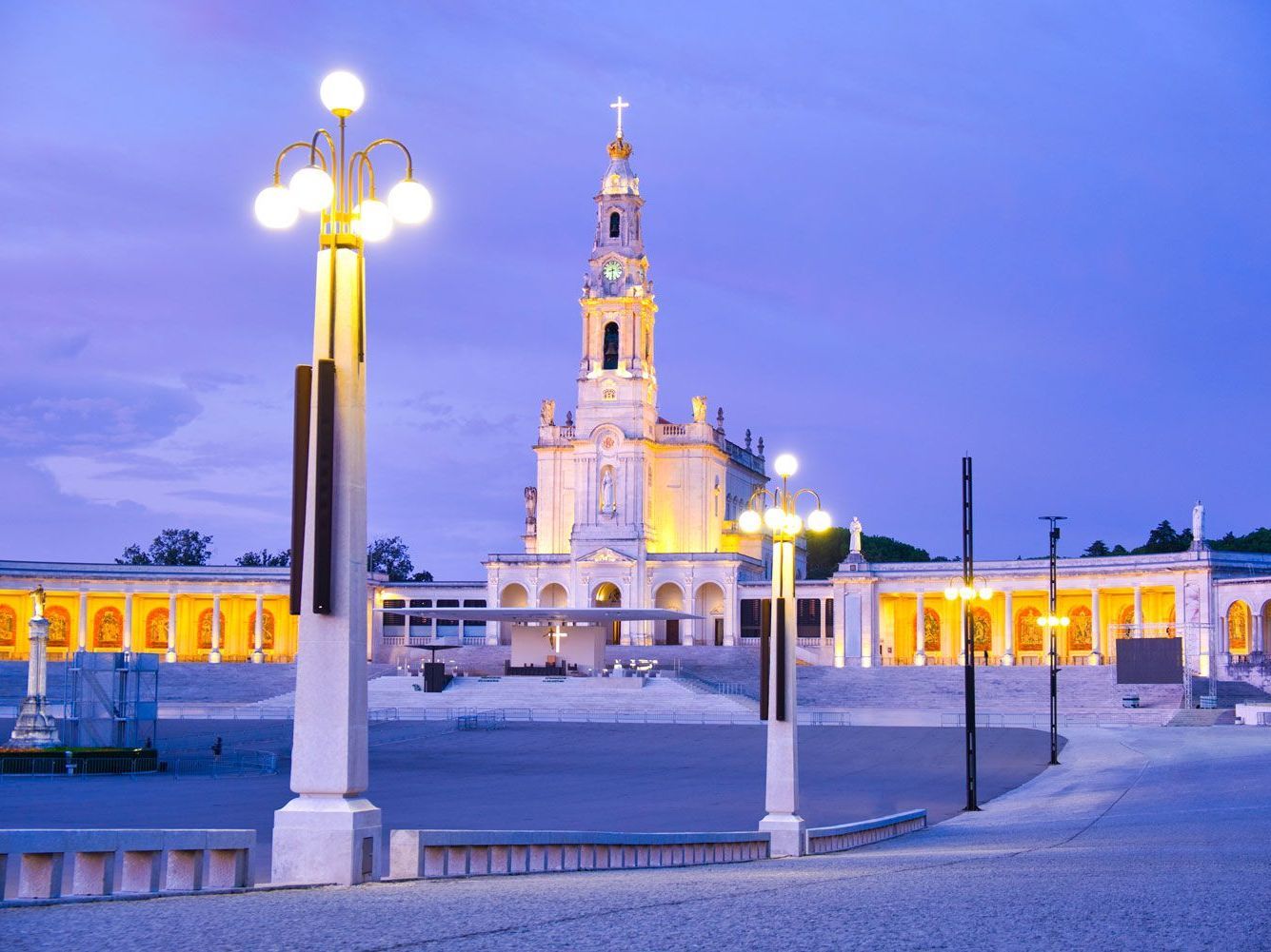 Basílica de Nuestra Señora del Rosario en Fátima, Portugal, al anochecer. Una iglesia alta y blanca con un campanario iluminado con luces doradas; una vasta plaza abierta.