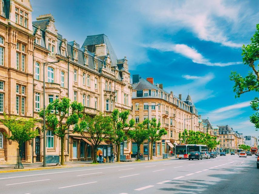 Una calle ancha bordeada de edificios ornamentados en una ciudad europea bajo un cielo azul brillante, con árboles, automóviles y peatones.