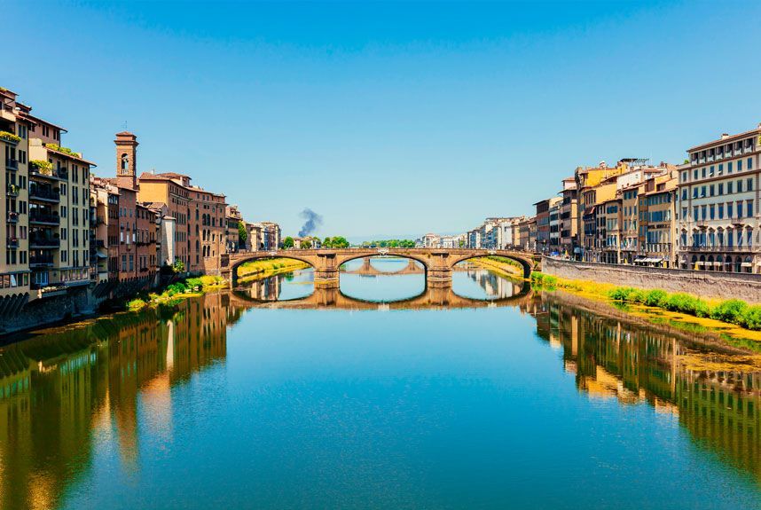 Una vista panorámica del puente Santa Trinita en Florencia, Italia, reflejado en el río Arno bajo un cielo azul claro.