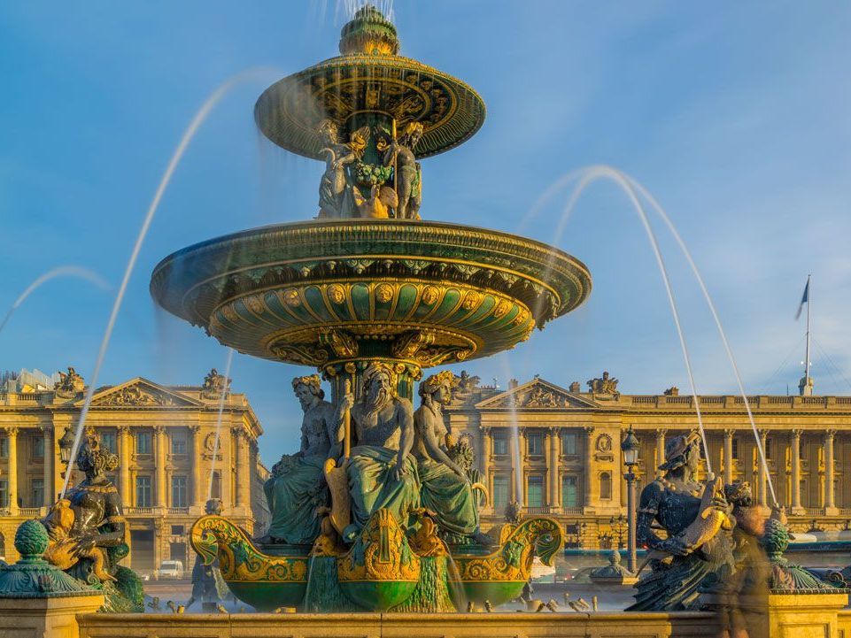Fuente en la Plaza de la Concordia, París, con figuras de bronce ornamentadas y surtidores. Día soleado, con edificios al fondo.