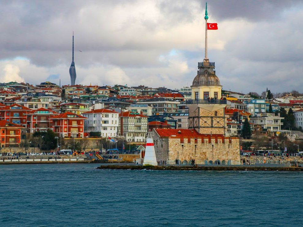 La Torre de la Doncella en Estambul, Turquía, con la bandera turca ondeando, frente a un fondo de edificios coloridos y un cielo nublado.