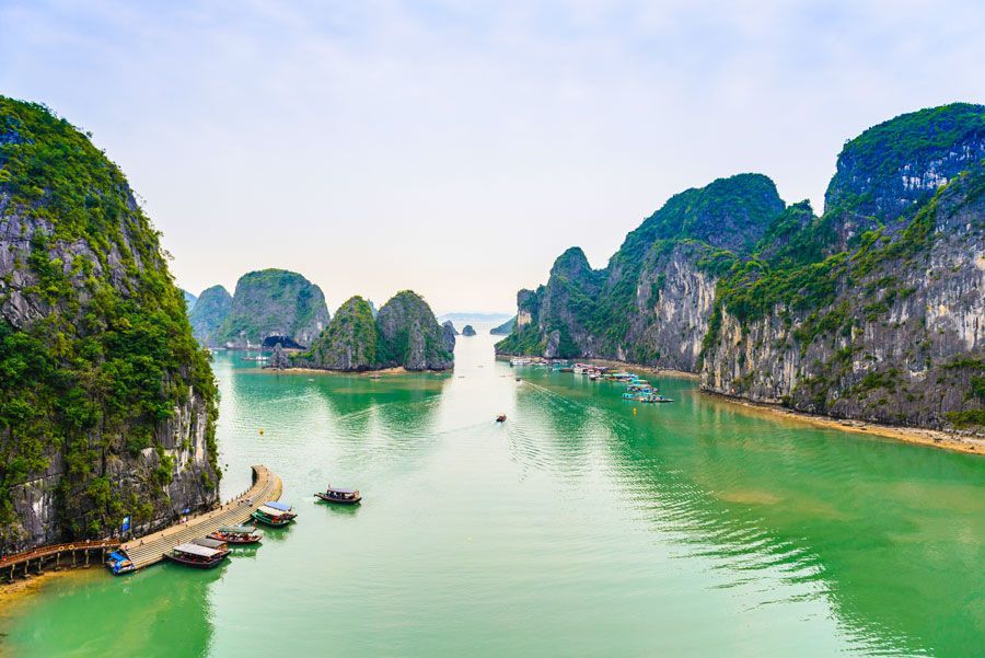 El agua verde rodea los karsts de piedra caliza en la bahía de Ha Long, Vietnam. Los barcos salpican el agua bajo un cielo nublado.