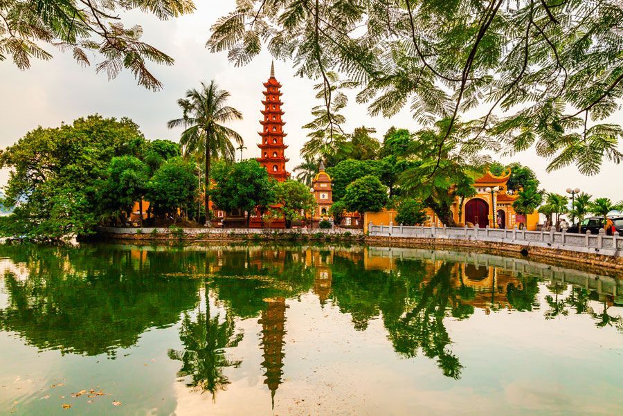 Una pagoda y una exuberante vegetación se reflejan en las tranquilas aguas. Un puente conduce al templo, enmarcado por frondosas ramas.