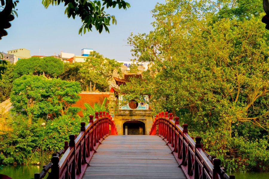 Puente de arco rojo sobre el agua, que conduce a un edificio tradicional enmarcado por exuberantes árboles verdes y un cielo azul brillante en Hanoi.