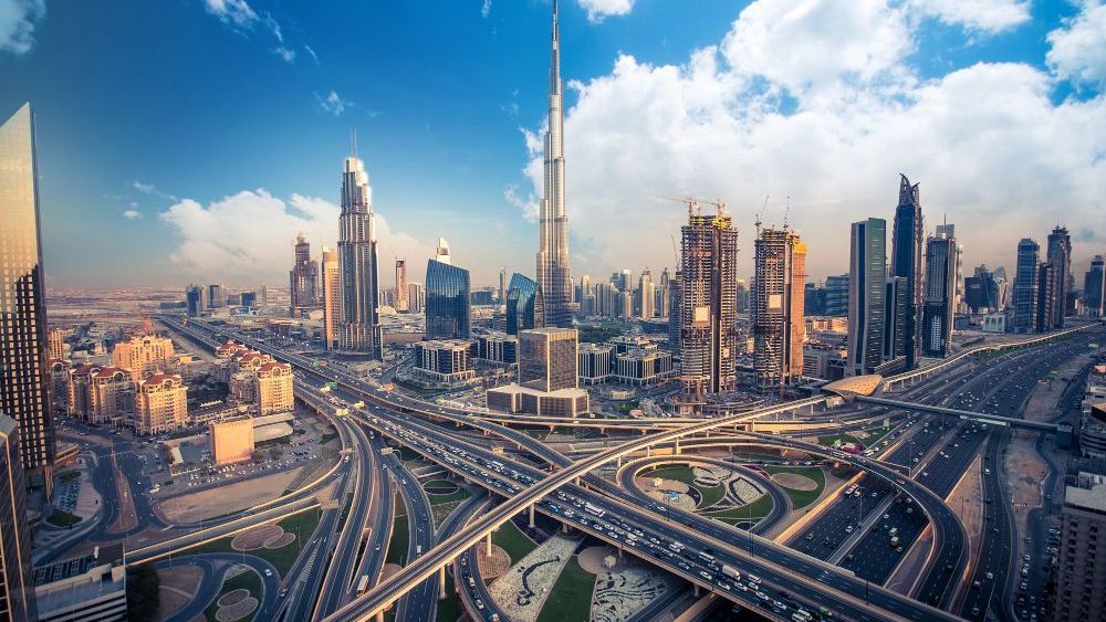 Un extenso paisaje urbano de Dubai, con el Burj Khalifa elevándose sobre otros rascacielos modernos, entrelazado por una compleja red de carreteras bajo un cielo azul.