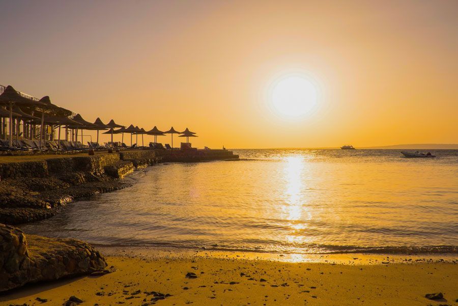 Puesta de sol dorada sobre una playa con sombrillas, tumbonas y un pequeño barco en el agua.