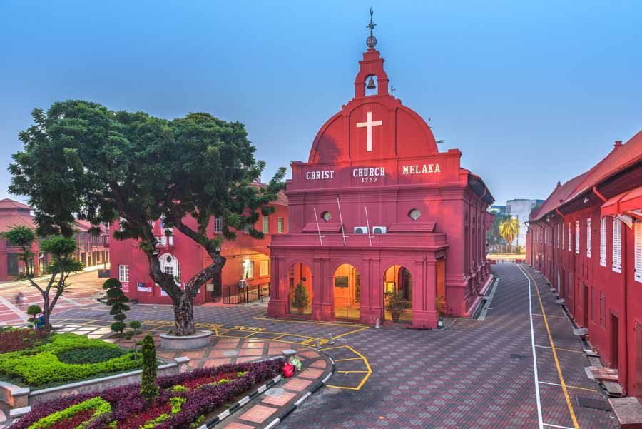 Iglesia de Cristo Rojo en Melaka, Malasia, con una gran cruz blanca y un campanario, rodeada de edificios rojos y exuberante vegetación.