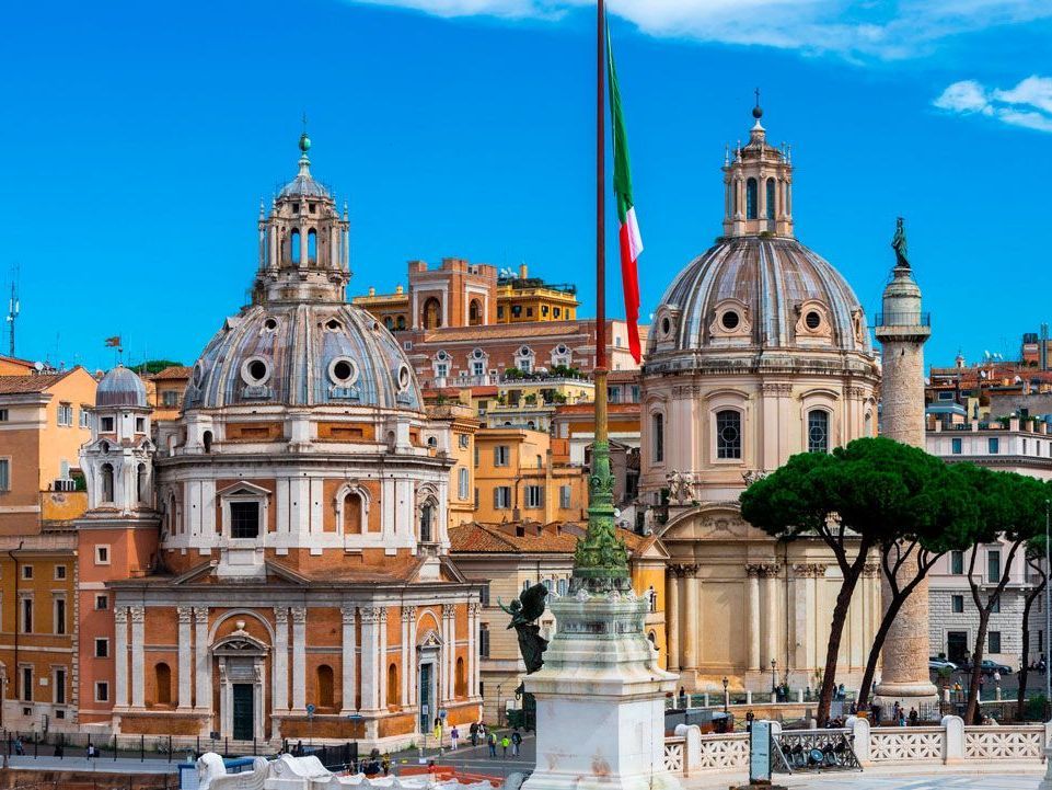 Una toma vibrante de edificios romanos con cúpulas ornamentadas contra un cielo azul claro, con la bandera italiana ondeando en primer plano.