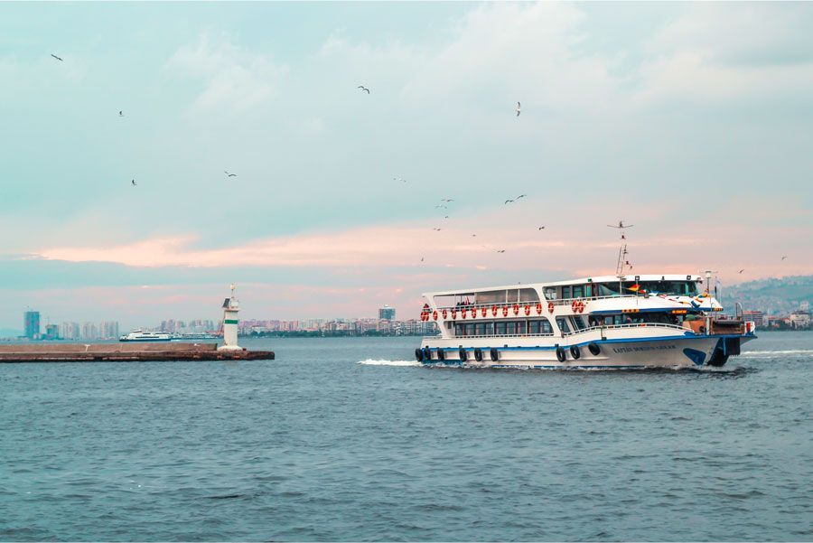 Un barco blanco y azul navega por un mar azul hacia un faro, con el horizonte de una ciudad al fondo.
