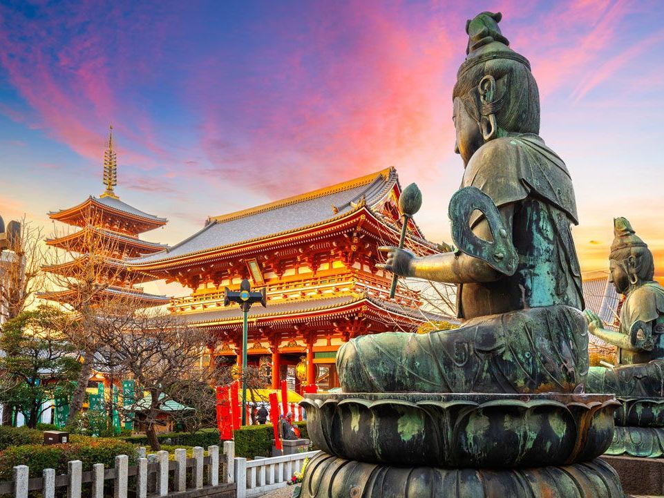 Una gran estatua de bronce de Buda se encuentra frente a un templo japonés tradicional con una pagoda de cinco pisos, bajo una colorida puesta de sol.