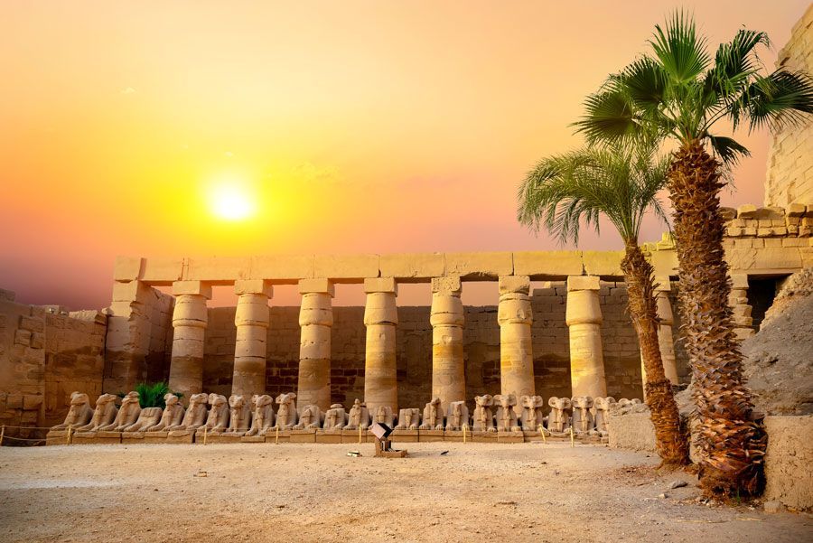 Una puesta de sol sobre el antiguo templo de Karnak en Luxor, Egipto, con imponentes columnas, estatuas y palmeras.