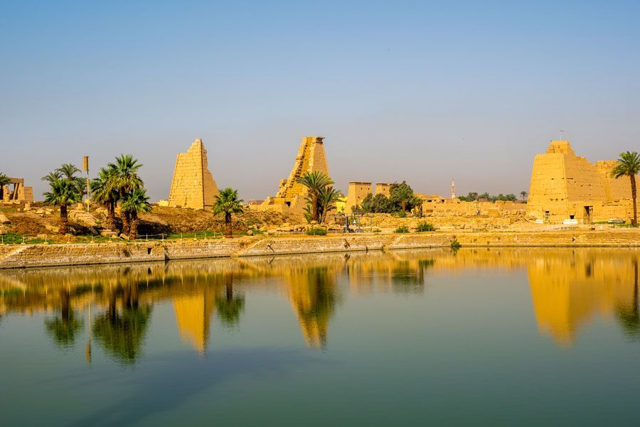 Ruinas de un antiguo templo reflejadas en un tranquilo lago azul, con palmeras y un cielo azul claro en el fondo.