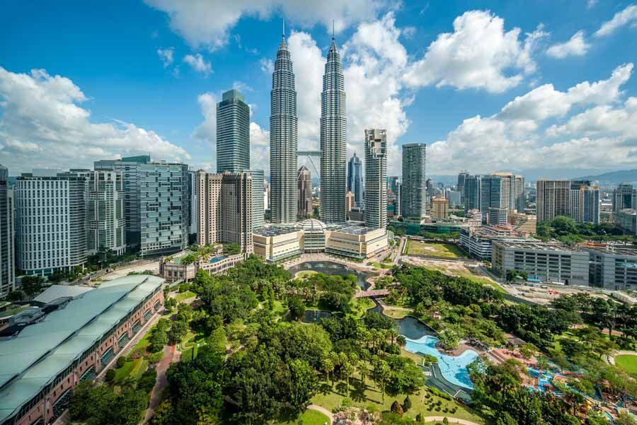 Las Torres Gemelas Petronas y otros rascacielos dominan el horizonte de Kuala Lumpur con un parque en primer plano, bajo un cielo azul brillante con nubes hinchadas.