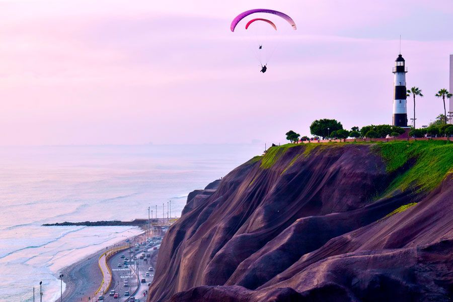 Parapentes sobrevuelan un acantilado costero en Lima, Perú, cerca de un faro rayado. Abajo se divisan el océano y una playa de arena.