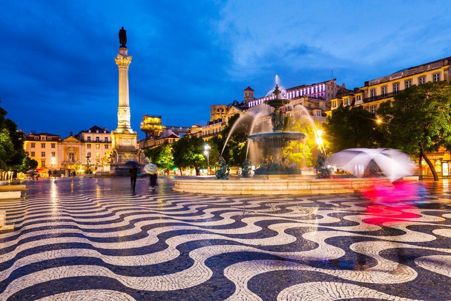 Una vista nocturna de la Plaza Rossio en Lisboa, Portugal, con una fuente, un pavimento estampado y una columna alta.