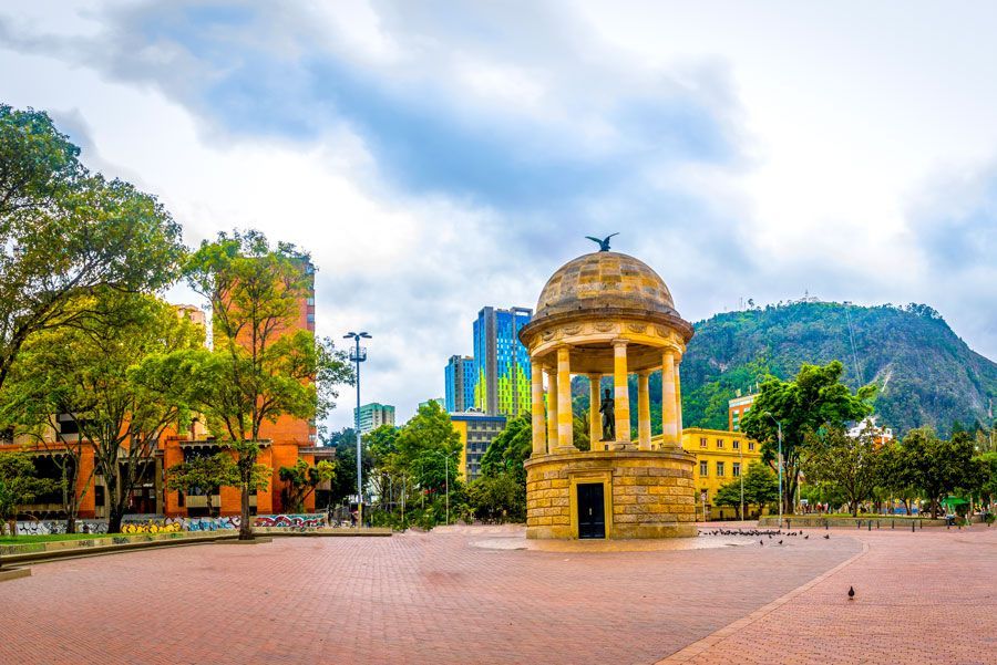 Plaza de Bolívar en Bogotá, Colombia. Un kiosco de piedra con una estatua se alza en una amplia plaza de pavimento rojo, rodeada de árboles y edificios bajo un cielo nublado.