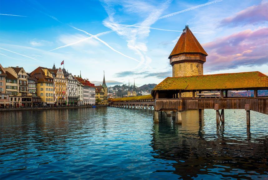 Puente de la capilla de madera en Lucerna, Suiza, con vista al agua y edificios frente al mar bajo un cielo azul.