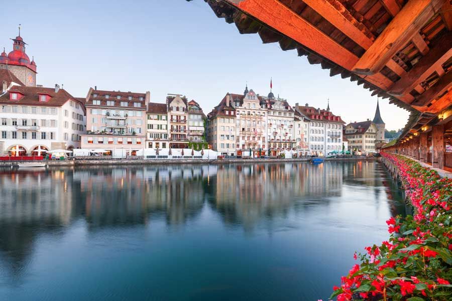 Puente de madera sobre un río tranquilo en Lucerna, Suiza. Los edificios bordean la orilla. Flores rojas y el puente de madera aparecen en primer plano.