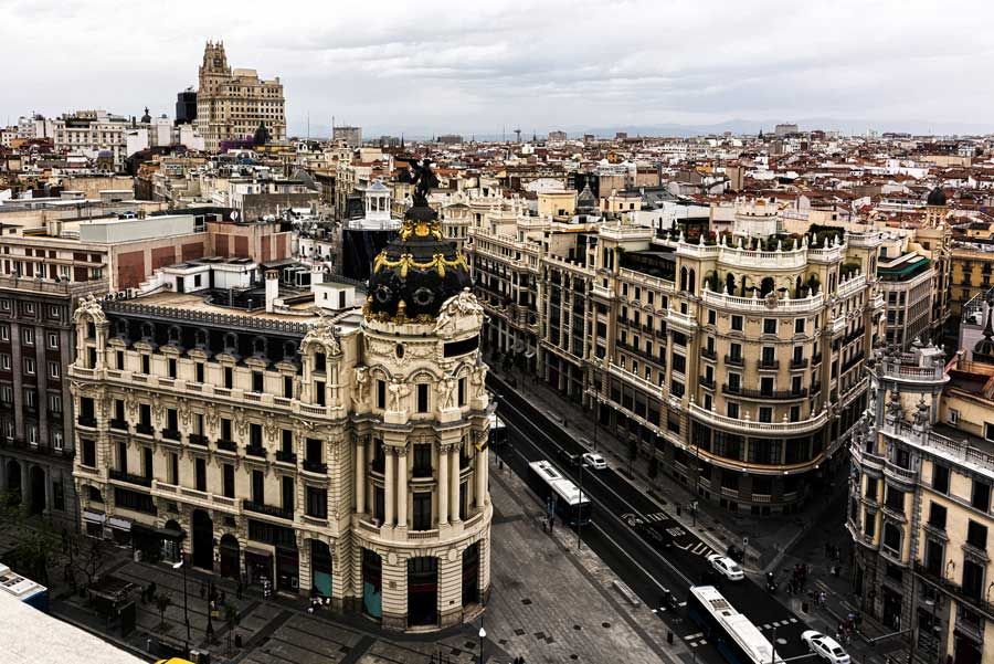 Paisaje urbano de Madrid, España, con edificios ornamentados de colores claros y una estructura abovedada en primer plano. Cielo gris en lo alto.