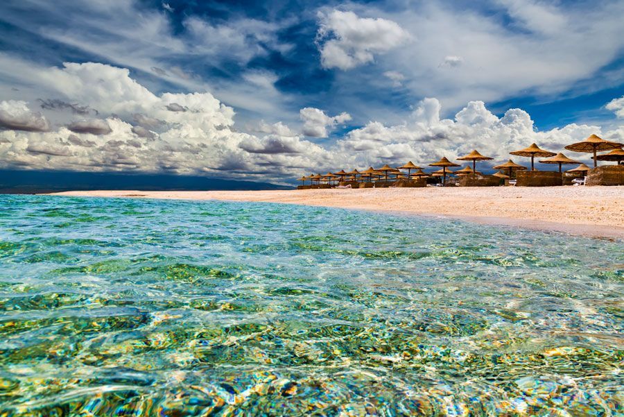 Agua azul baña una playa de arena bajo un cielo nublado, con sombrillas de playa al fondo.