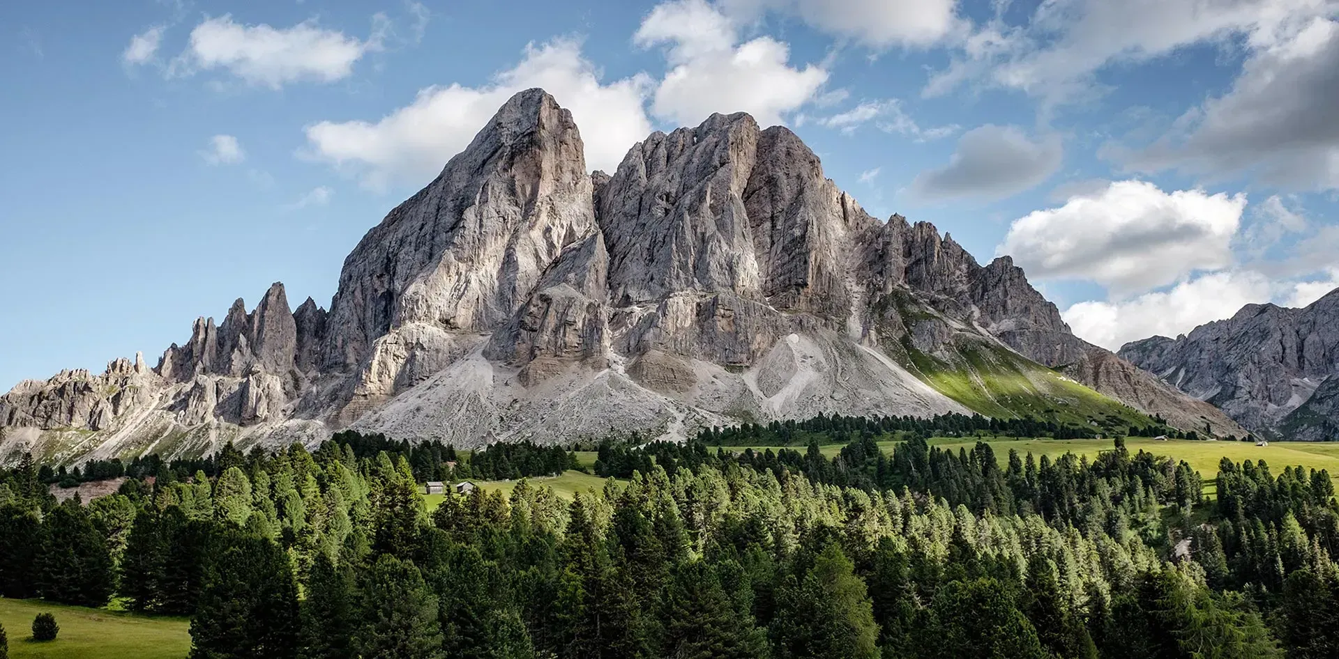 Una escarpada cadena montañosa se eleva detrás de un denso bosque de árboles verdes bajo un cielo azul con nubes dispersas.