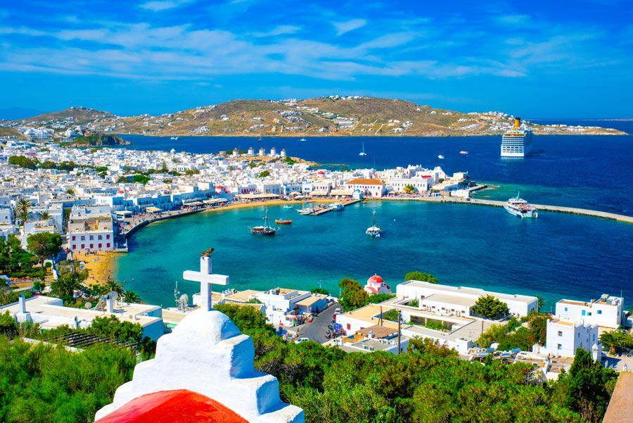Vista panorámica de Mykonos, Grecia, con edificios blancos que bordean una bahía azul, una cruz prominente en primer plano y una montaña en el fondo.