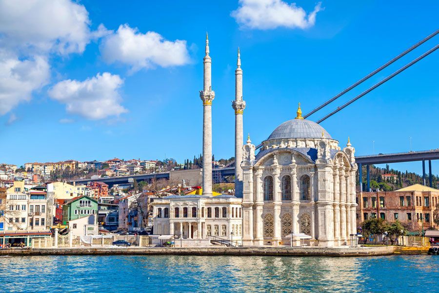 Una mezquita blanca con dos minaretes se encuentra frente al mar en Estambul, Turquía, con un puente al fondo bajo un cielo azul.