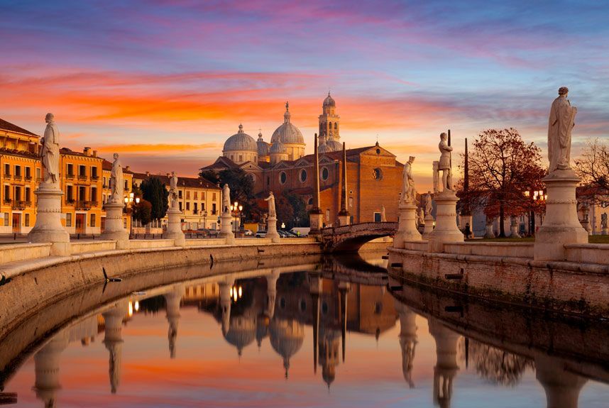 Canal del río en Padua, Italia, bordeado de estatuas bajo un cielo de atardecer vibrante que se refleja en el agua; edificios y una iglesia con cúpula en el fondo.