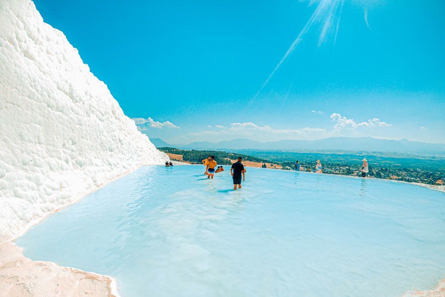 Gente chapoteando en las piscinas termales de color azul claro de Pamukkale, Turquía, contra terrazas de travertino blanco y un cielo azul claro.