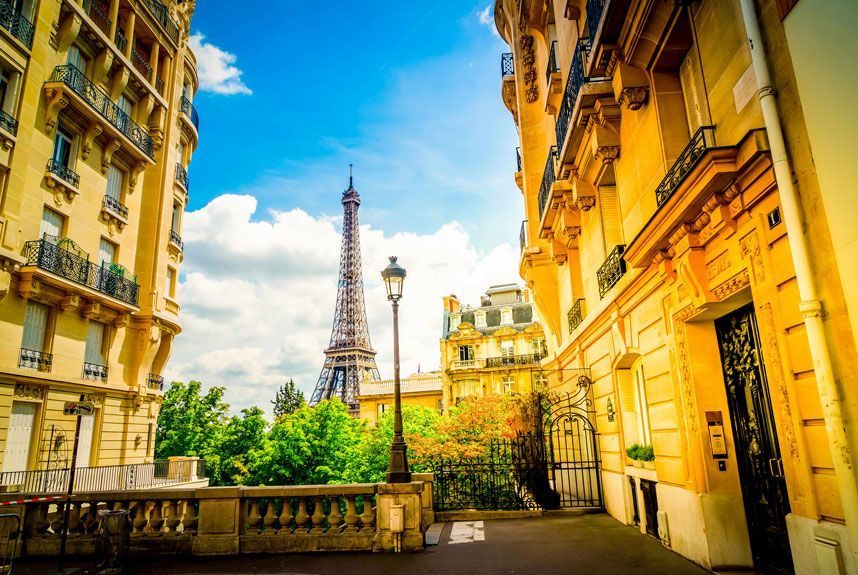 Torre Eiffel enmarcada por edificios parisinos en un día soleado, con una farola y follaje en primer plano.