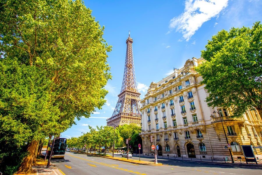 Torre Eiffel en París, Francia, visible detrás de una calle bordeada de árboles y edificios ornamentados bajo un cielo azul soleado.