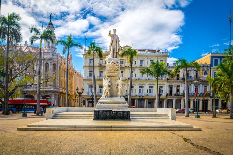 Estatua de José Martí en La Habana, Cuba, en una plaza con edificios coloniales, palmeras y cielo azul.
