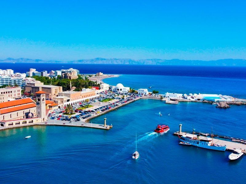 Vista aérea del puerto de Kos, Grecia, con agua azul, edificios, barcos y un cielo despejado.