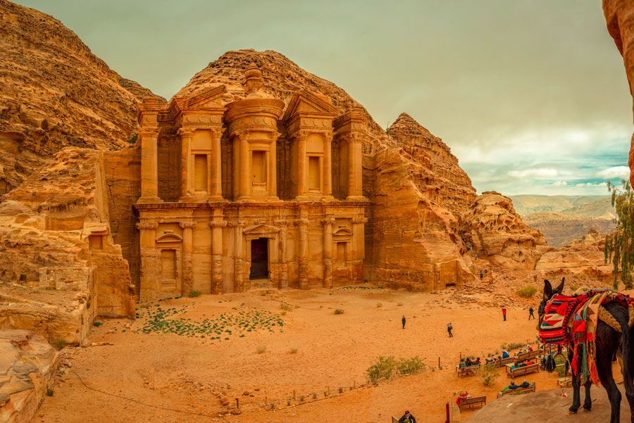 Un gran edificio antiguo tallado en una pared de arenisca en Petra, Jordania, con gente y un burro cerca.