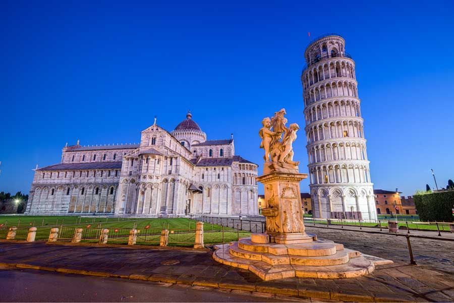 Pisa, Italia, al atardecer: La Torre Inclinada de Pisa junto a la Catedral de Pisa. Una estatua en primer plano está iluminada por una cálida luz.