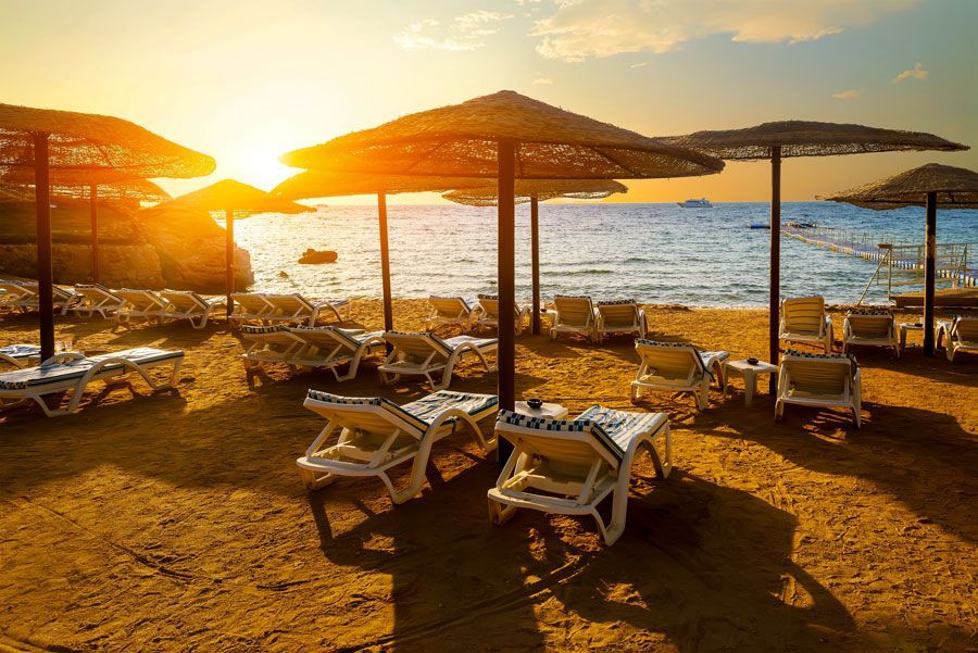 Escena de playa al atardecer con tumbonas bajo sombrillas de paja. El sol brilla sobre el agua y la arena.