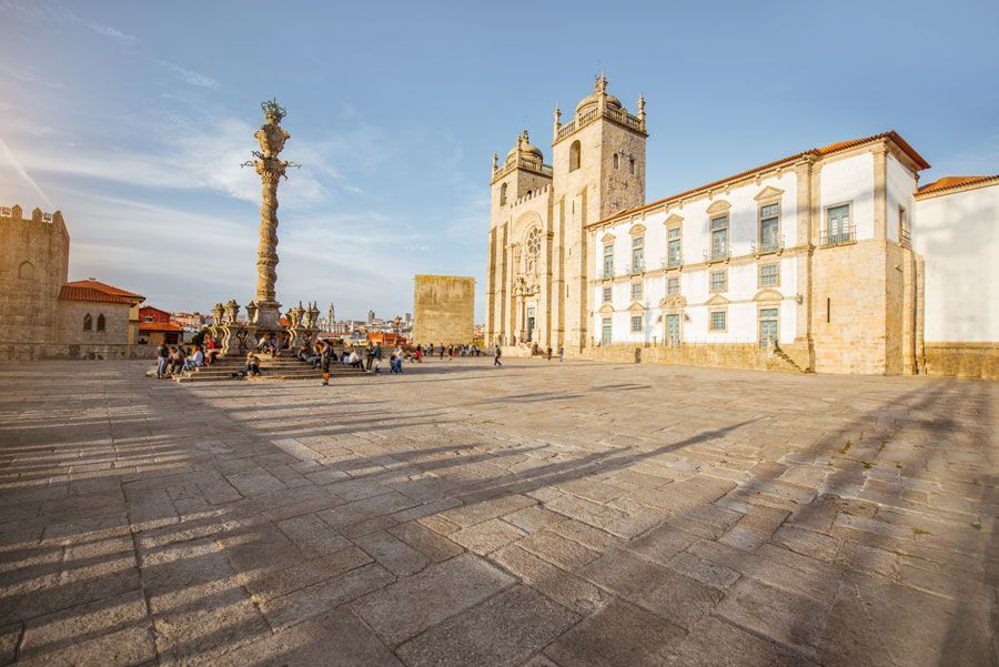 Una plaza soleada en Oporto, Portugal, con una catedral, una columna y gente paseando. Edificios de piedra y un cielo azul dominan la escena.