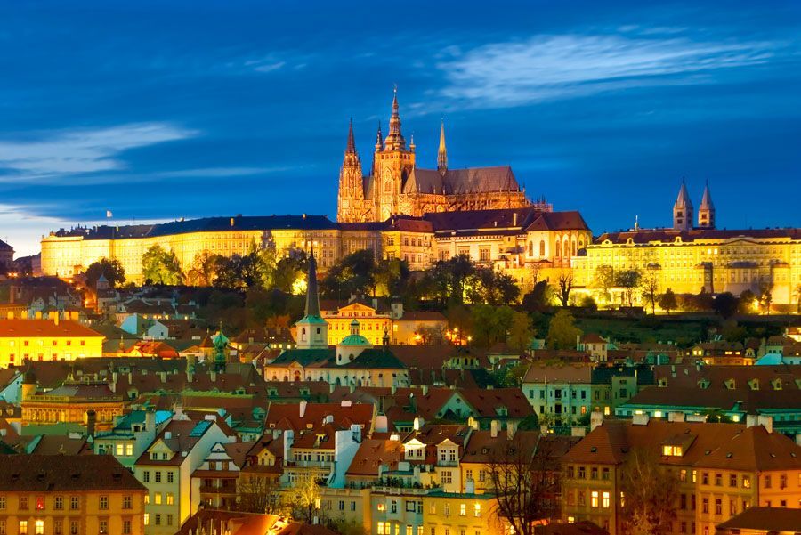 Complejo del Castillo de Praga iluminado al anochecer, con vistas a un colorido paisaje urbano con tejados y edificios.