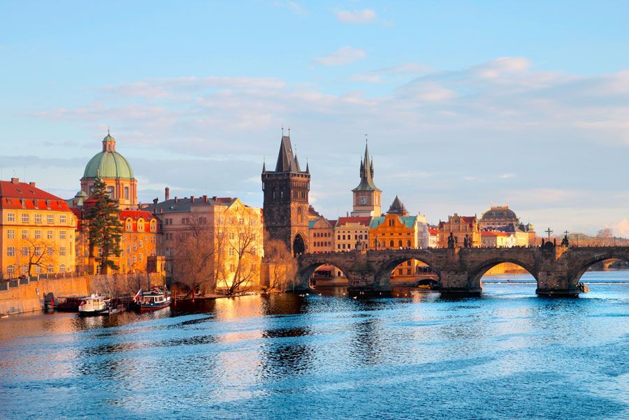 El horizonte de Praga al amanecer: edificios históricos, puente de piedra y barcos reflejados en el tranquilo río.