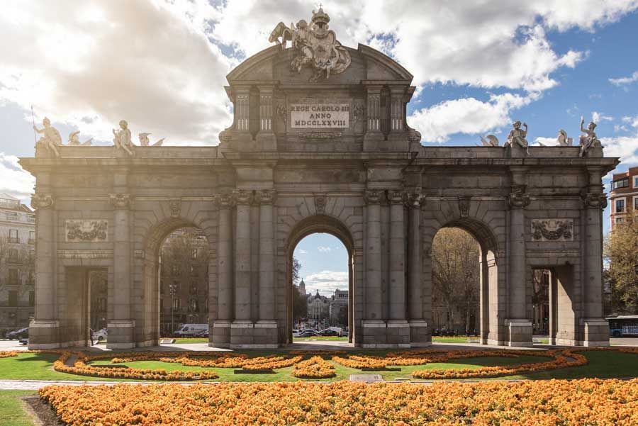 La Puerta de Alcalá, Madrid, un gran arco de piedra con columnas y esculturas bajo un cielo nublado. Flores anaranjadas rodean la base.