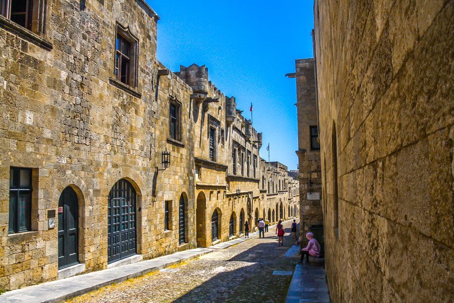 Calle adoquinada bordeada de antiguos edificios de piedra en Rodas, Grecia. Varias personas caminan o se sientan al sol.