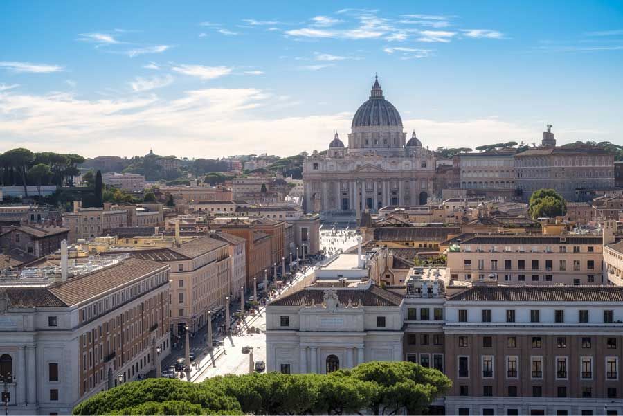 Vista de Roma, Italia, con la Basílica de San Pedro dominando el horizonte bajo un cielo azul brillante. Edificios y carreteras ocupan el primer plano.